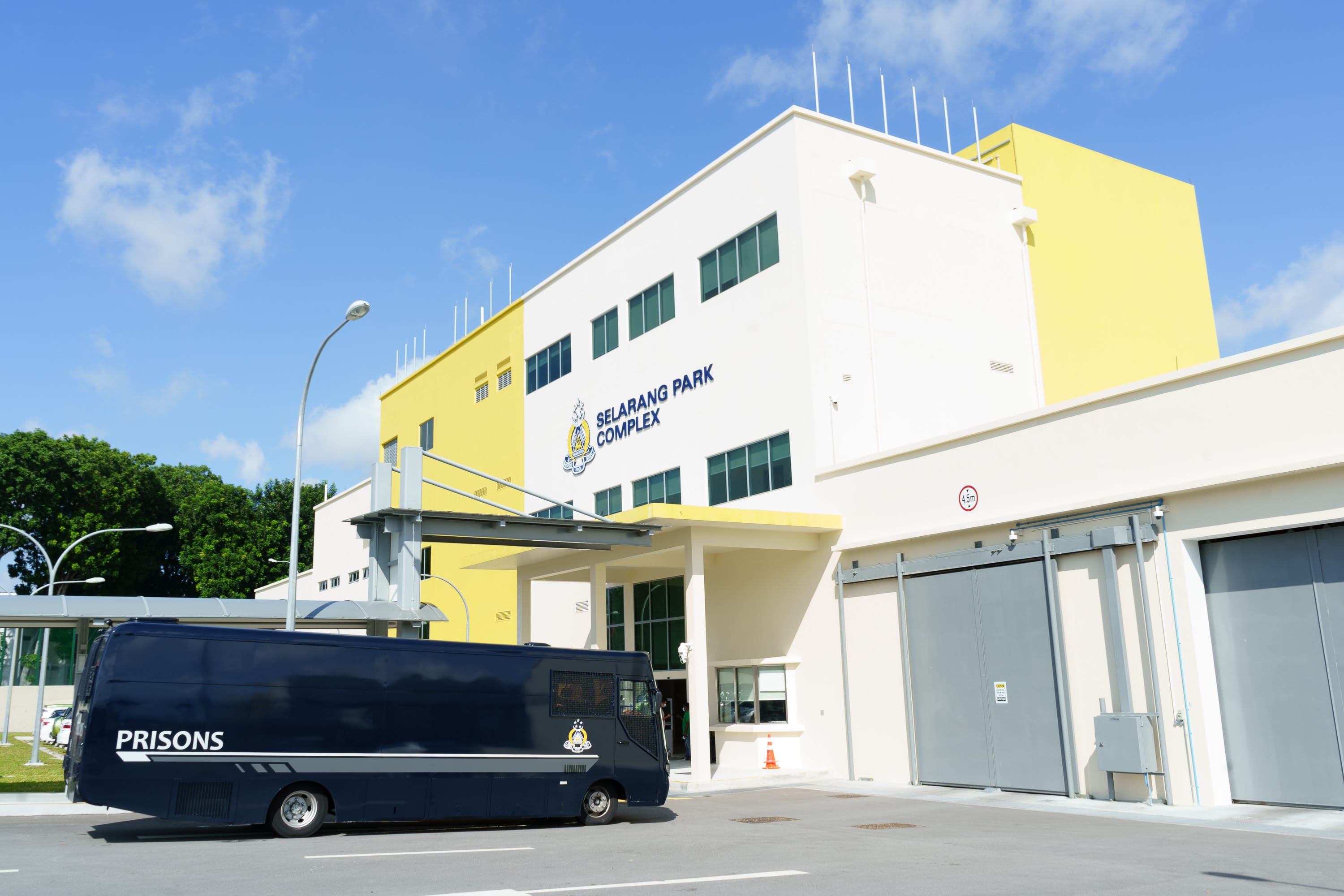 A dark blue prison bus is parked in front of a white and yellow building labeled "Selarang Park Complex."